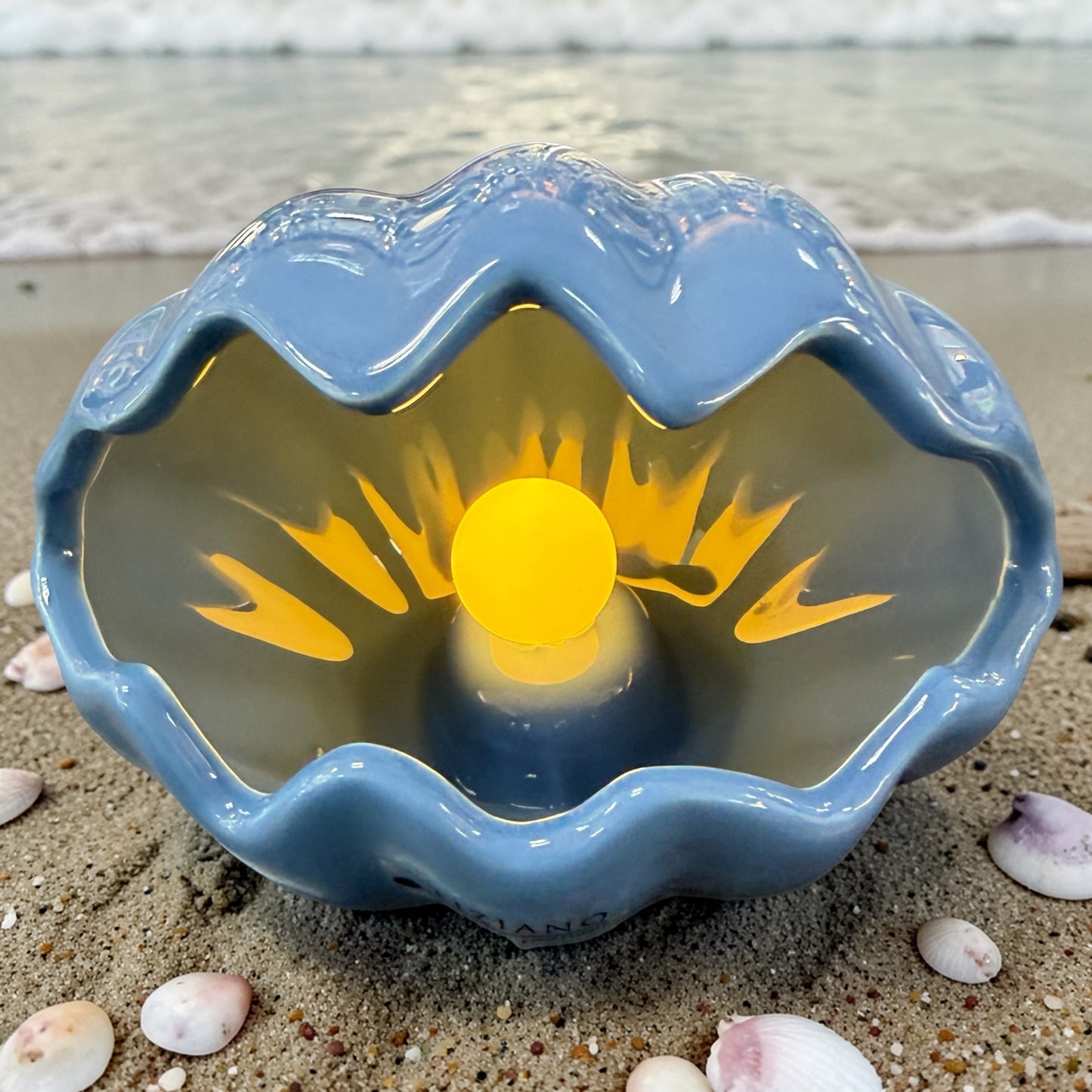 Eine blaue Keramik-Muschel mit leuchtender LED-Perle steht dekorativ am Strand, umgeben von Sand und kleinen Muscheln.