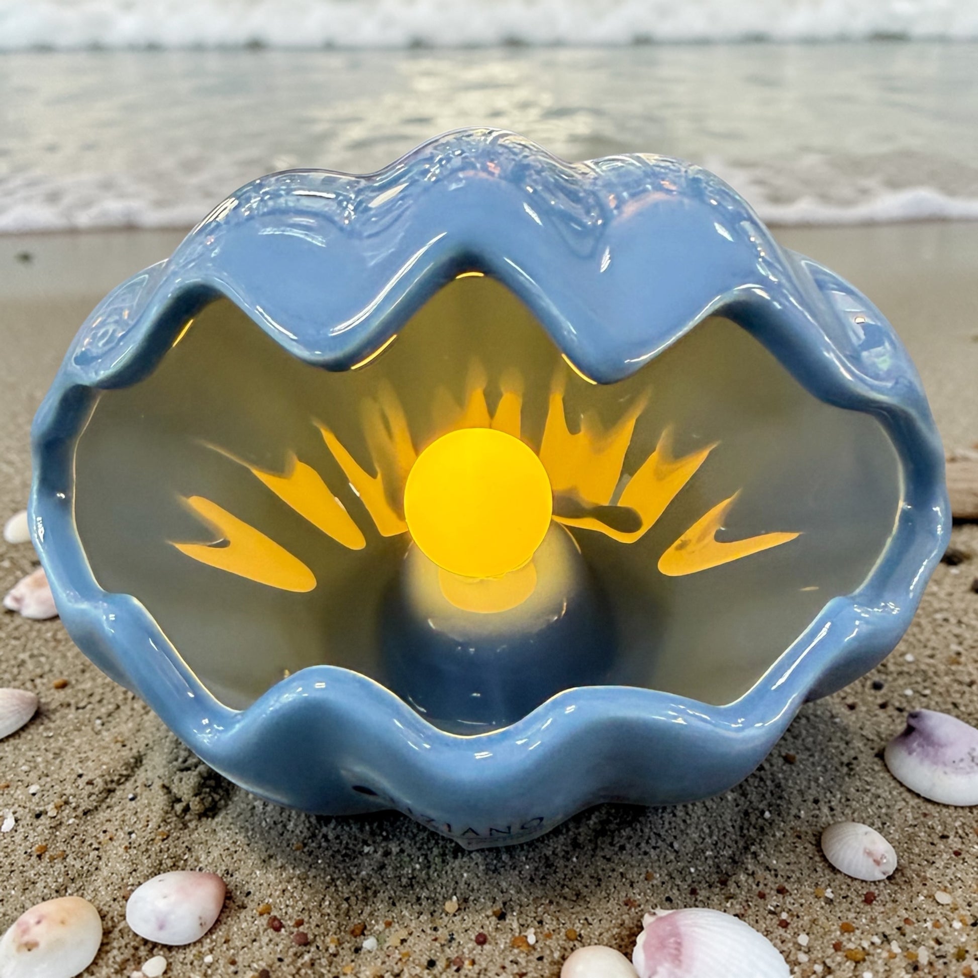 Eine blaue Keramik-Muschel mit leuchtender LED-Perle steht dekorativ am Strand, umgeben von Sand und kleinen Muscheln.