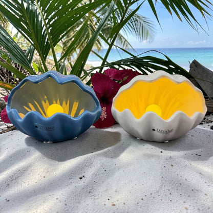 Zwei dekorative Keramik-Muscheln mit LED-Perle in Blau und Weiß stehen im Sand vor tropischer Strandkulisse mit Palmen und Meer.
