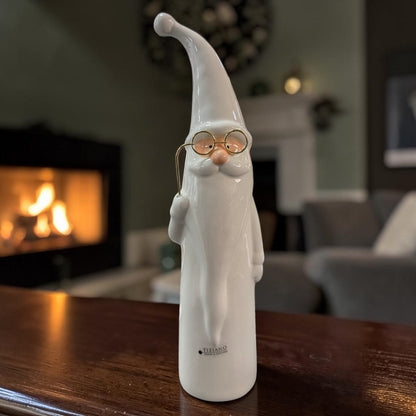 Cremeweißer Tiziano Keramik-Nikolaus mit langer Mütze, Bart und goldfarbener Brille auf einem dunklen Holztisch vor einem Kamin.