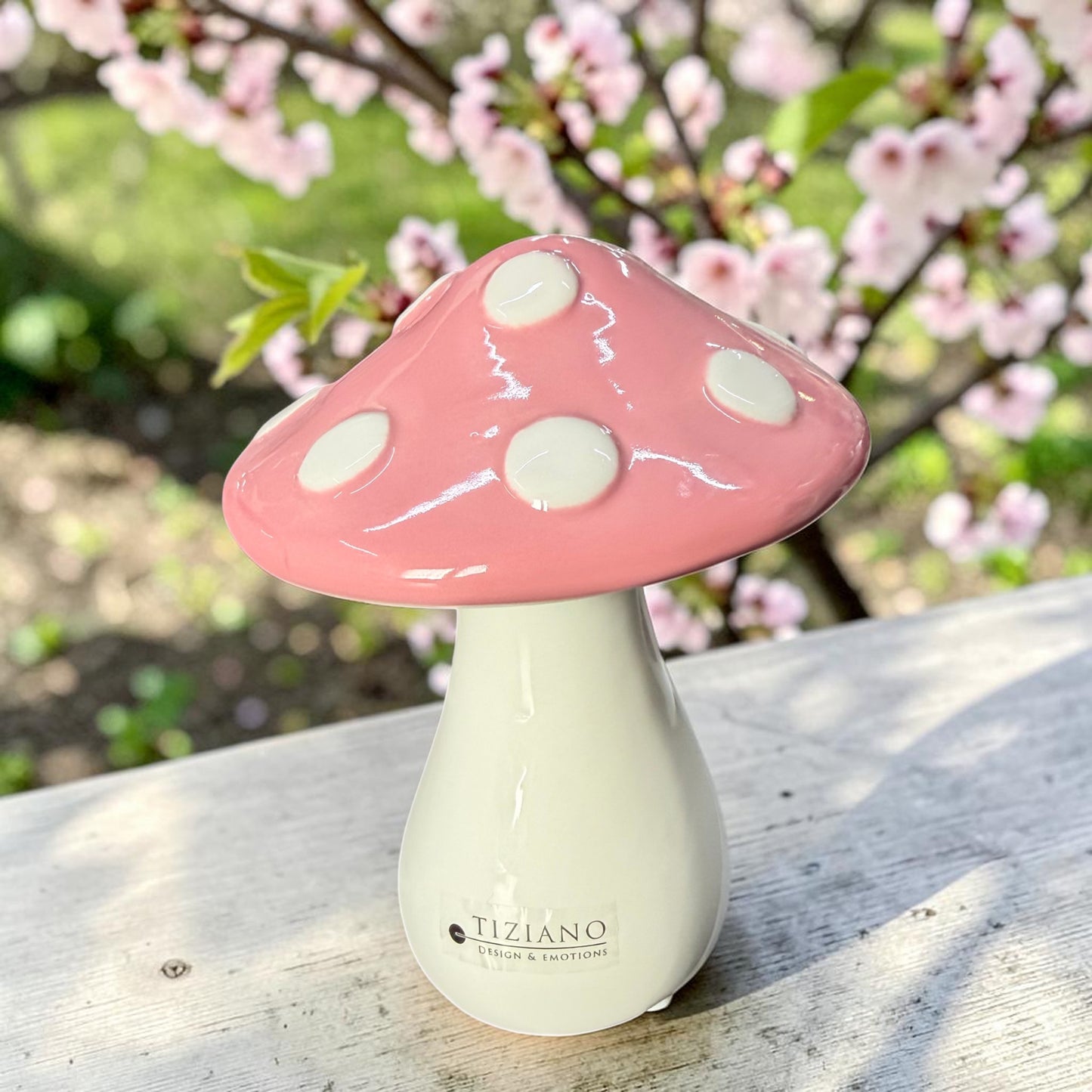 Tiziano Keramik-Pilz Ponti mit weißem Stiel und rosafarbenem Hut mit weißen Punkten auf einer grauen Holzfläche vor rosa Blüten.