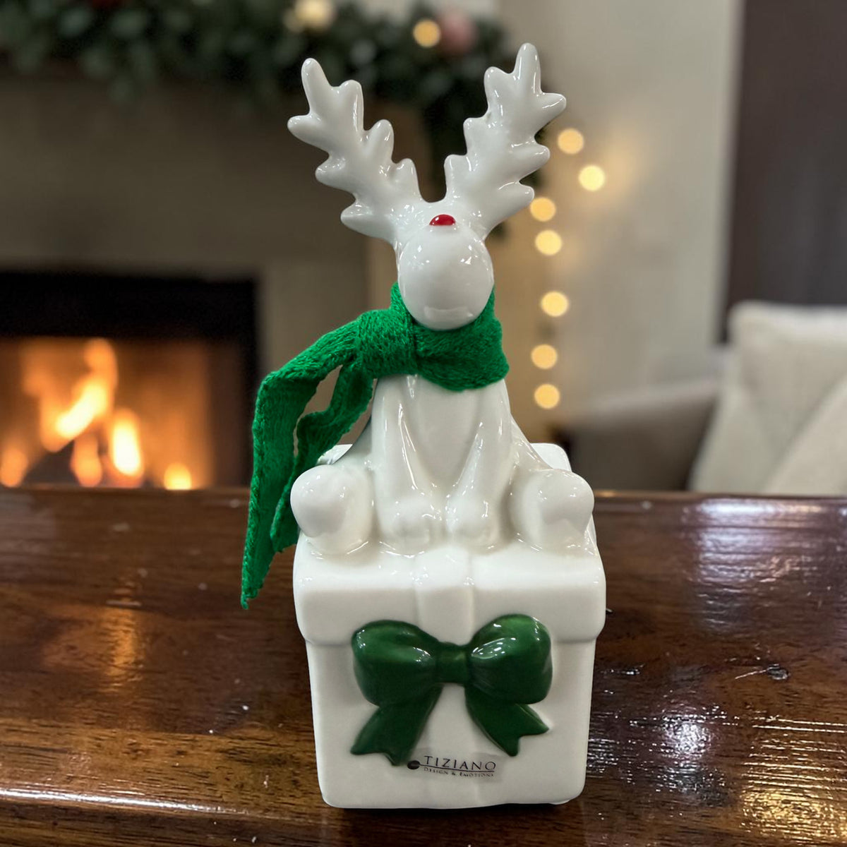 Cremeweißes Tiziano Keramik-Rentier mit grünem Schal auf einem weißen Geschenk mit grüner Schleife vor einem Kamin.