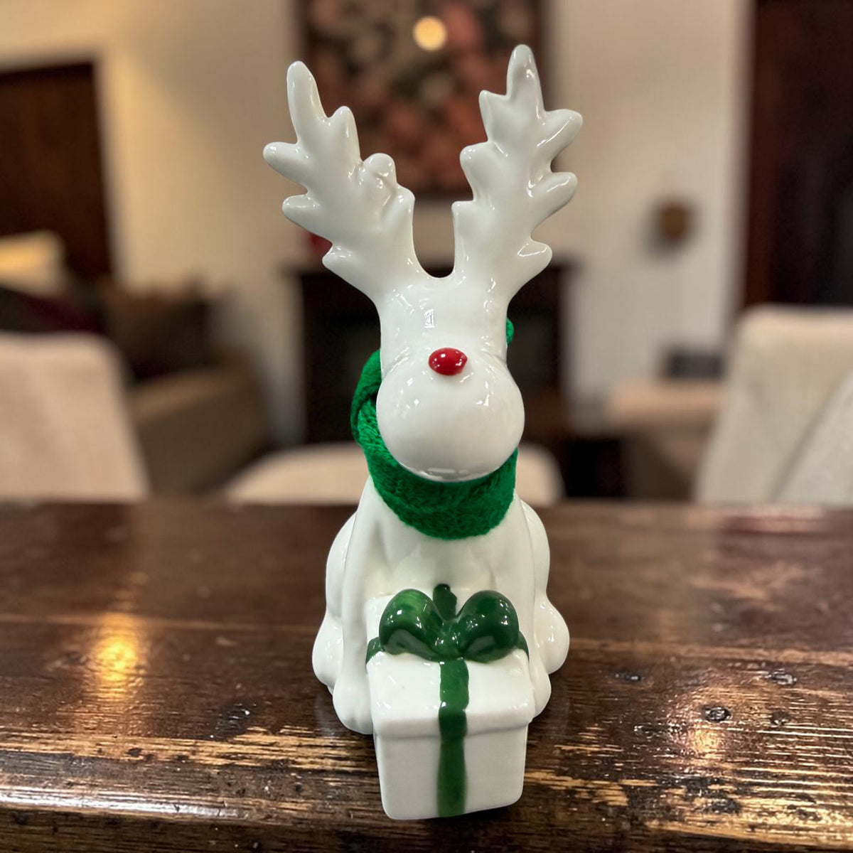 Cremeweißes Tiziano Keramik-Rentier mit grünem Schal und weißem Geschenk mit grüner Schleife vor Tannengrün und warmweißer Lichterkette.