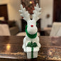 Cremeweißes Tiziano Keramik-Rentier mit grünem Schal und weißem Geschenk mit grüner Schleife vor Tannengrün und warmweißer Lichterkette.