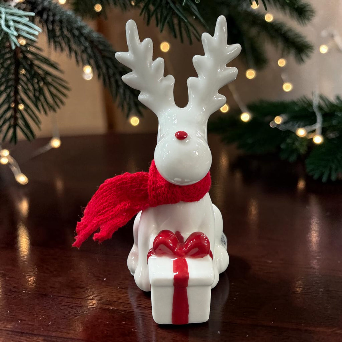 Cremeweißes Tiziano Keramik-Rentier mit rotem Schal und weißem Geschenk mit roter Schleife vor Tannengrün und warmweißer Lichterkette.