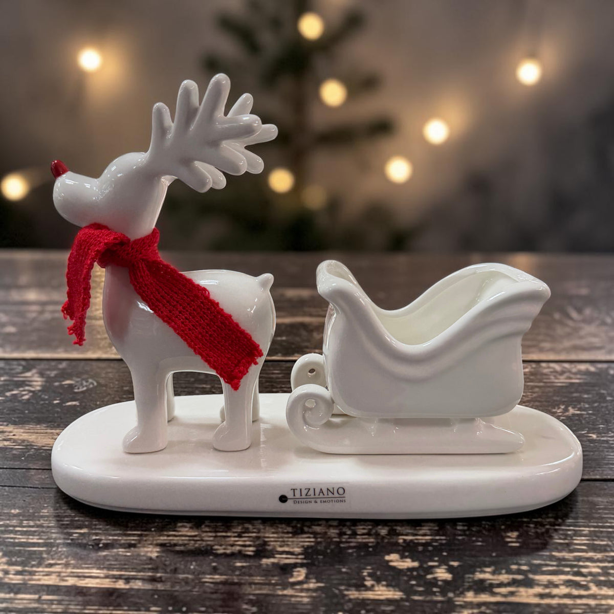 Tiziano Keramik Rentier in Cremeweiß mit rotem Schal und Schlitten auf einem cremeweißen Sockel vor weihnachtlicher Beleuchtung.