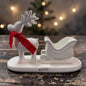 Tiziano Keramik Rentier in Cremeweiß mit rotem Schal und Schlitten auf einem cremeweißen Sockel vor weihnachtlicher Beleuchtung.