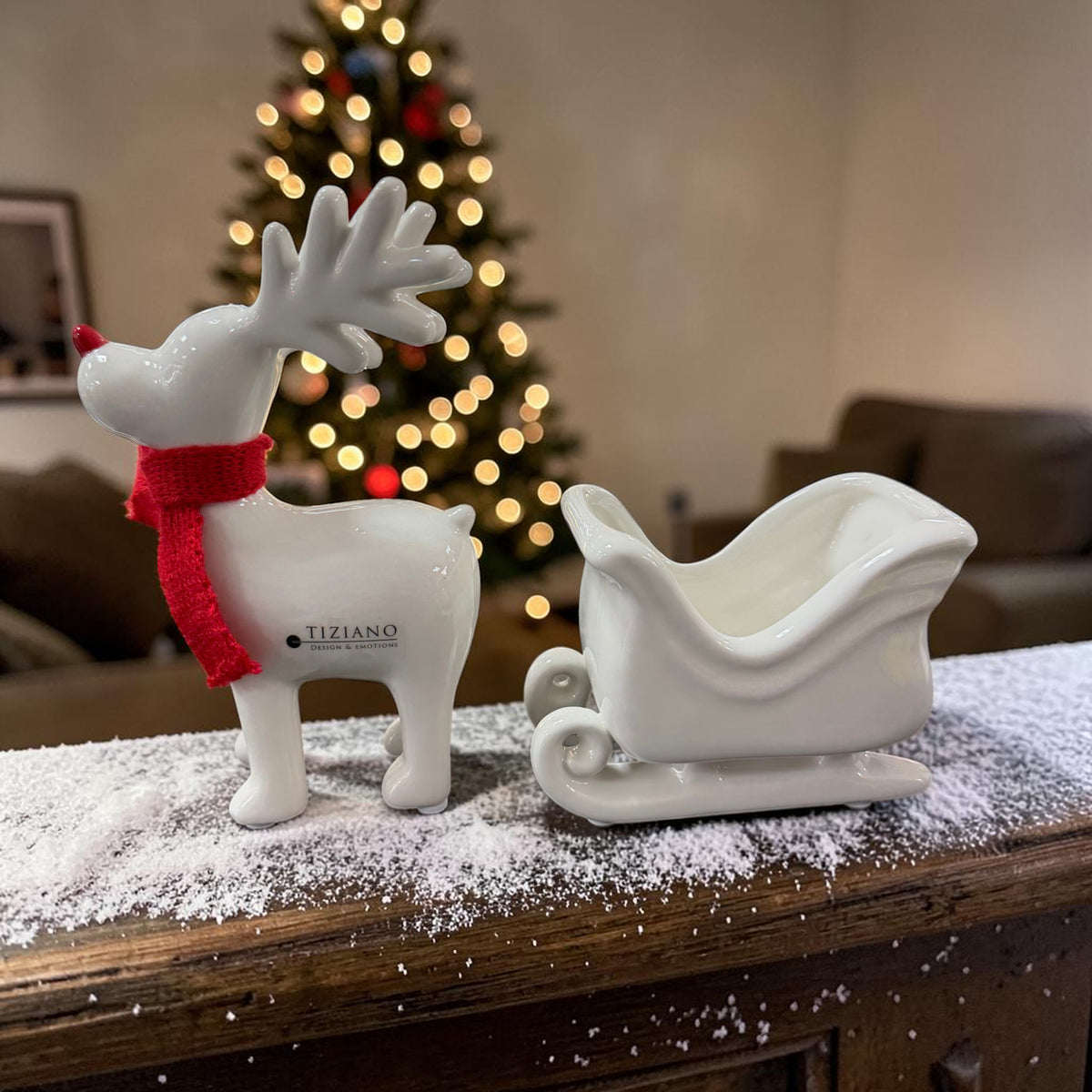 Tiziano Keramik Rentier in Cremeweiß mit rotem Schal neben einem cremeweißen Schlitten vor einem beleuchteten Weihnachtsbaum.