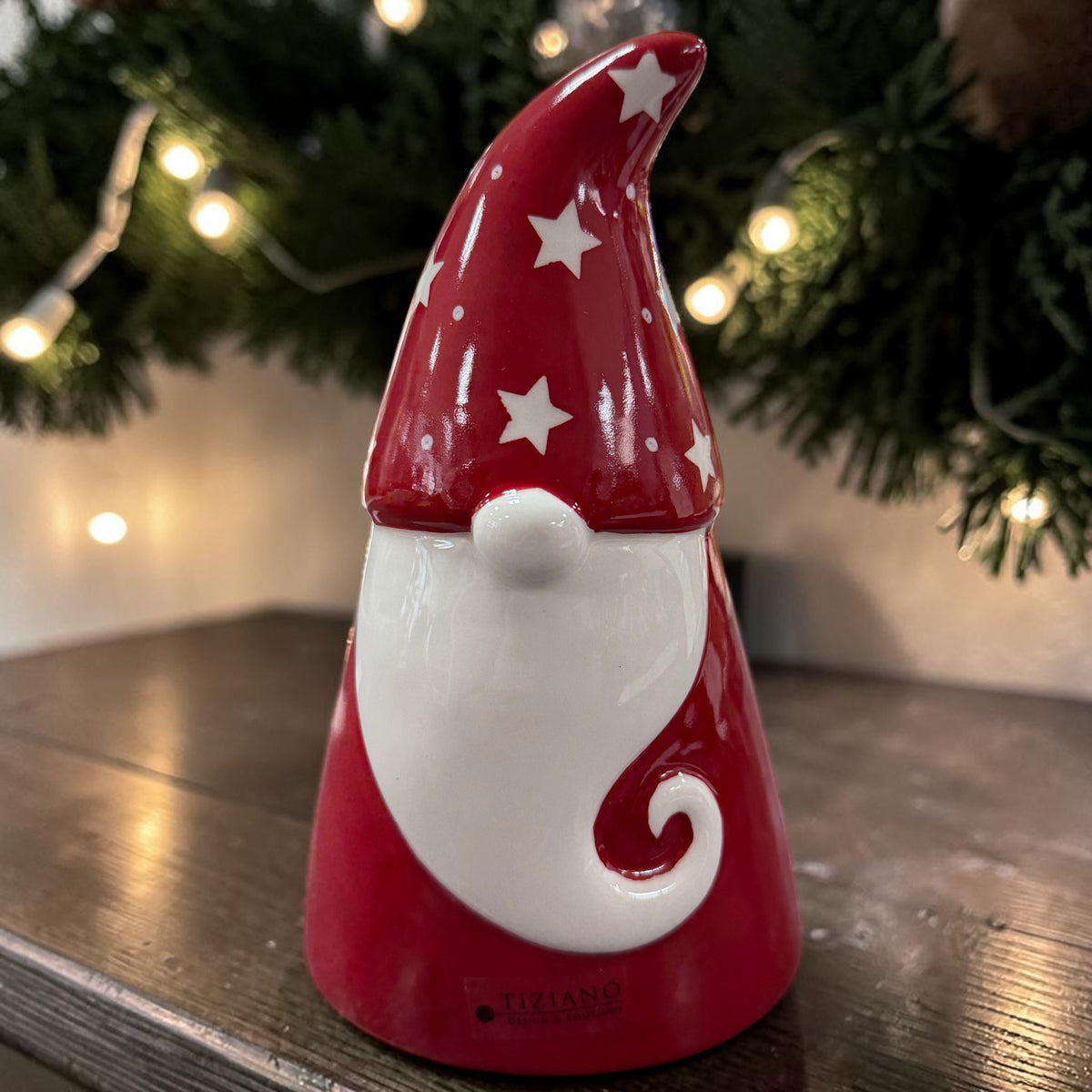 Roter Tiziano Keramik Weihnachtsmann mit weißem Bart und Sternenmütze vor Tannengrün und warm leuchtender Lichterkette.
