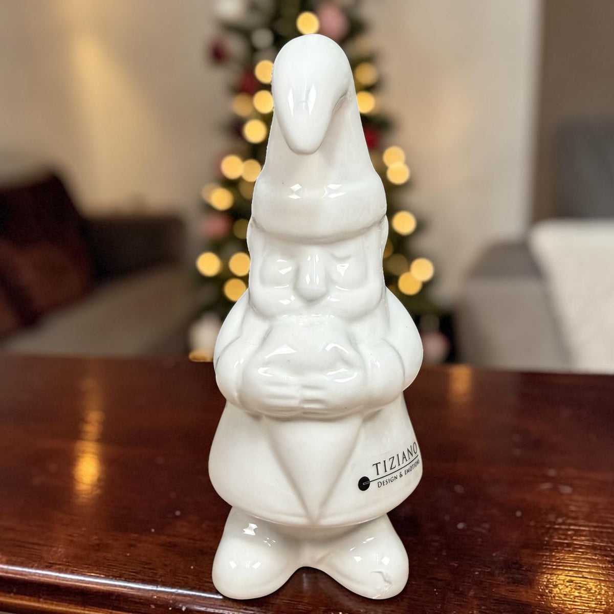 Großer cremeweißer Tiziano Keramik-Weihnachtsmann mit langer Zipfelmütze auf einem Holztisch vor einem beleuchteten Weihnachtsbaum.