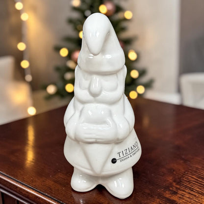 Kleiner cremeweißer Tiziano Keramik-Weihnachtsmann mit langer Zipfelmütze auf einem Holztisch vor einem beleuchteten Weihnachtsbaum.