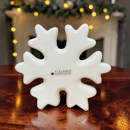 Große cremeweiße Tiziano Keramik-Schneeflocke auf einem Holztisch mit weihnachtlicher Beleuchtung im Hintergrund.