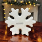 Große cremeweiße Tiziano Keramik-Schneeflocke auf einem Holztisch mit weihnachtlicher Beleuchtung im Hintergrund.