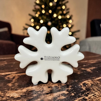 Kleine cremeweiße Tiziano Keramik-Schneeflocke auf einem Holztisch vor einem beleuchteten Weihnachtsbaum.