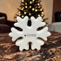 Kleine cremeweiße Tiziano Keramik-Schneeflocke auf einem Holztisch vor einem beleuchteten Weihnachtsbaum.