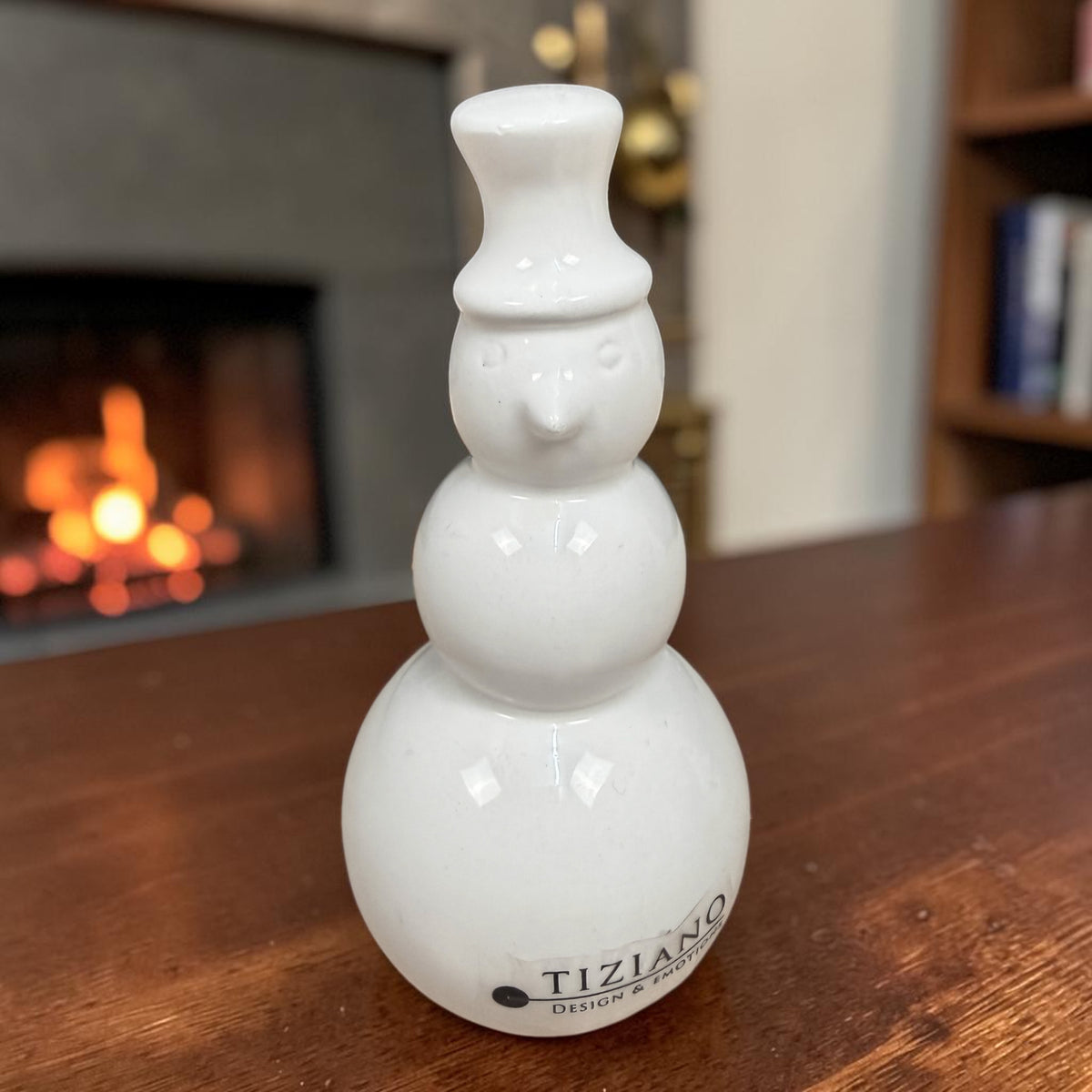 Cremeweißer Tiziano Keramik-Schneemann mit Zylinder auf einem Holztisch vor einem Kamin mit Feuer.
