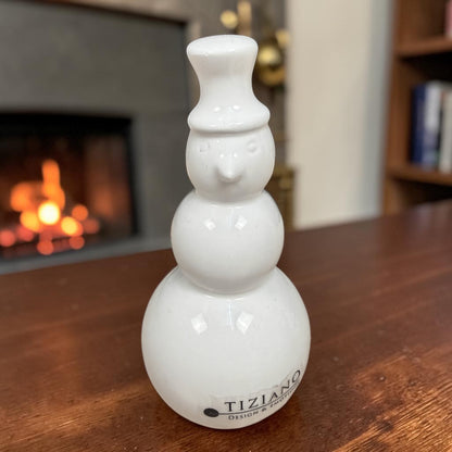 Cremeweißer Tiziano Keramik-Schneemann mit Zylinder auf einem Holztisch vor einem Kamin mit Feuer.