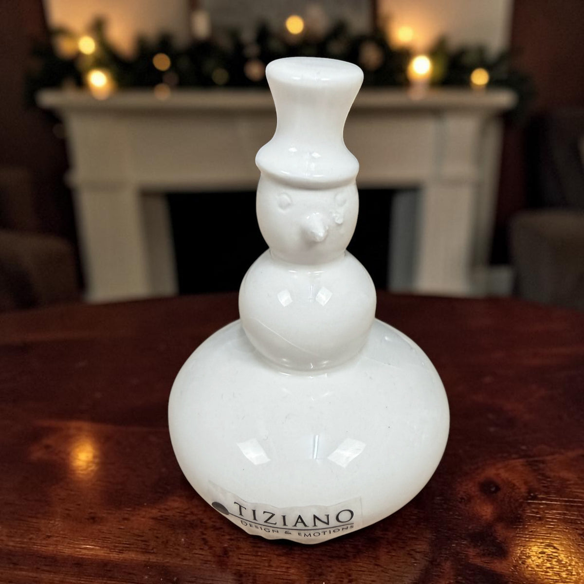 Kleiner cremeweißer Tiziano Keramik-Schneemann mit Zylinder auf einem Holztisch vor weihnachtlich beleuchtetem Kamin.