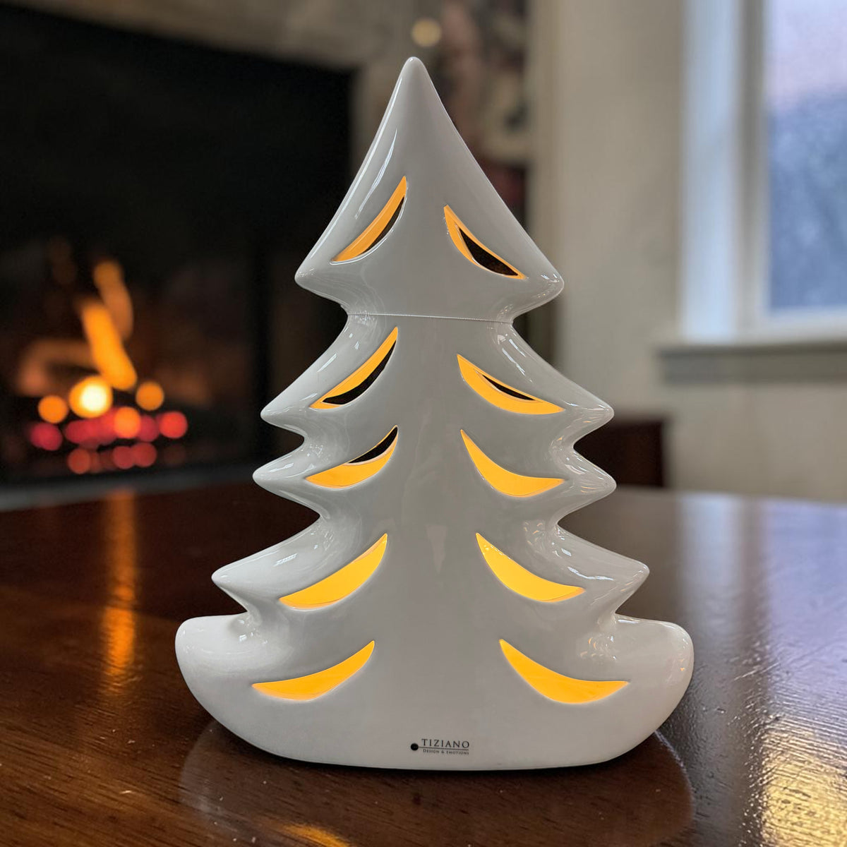 Cremeweißer Tiziano Keramik Tannenbaum mit warm beleuchteten Durchbrüchen auf einem Holztisch vor einem Kamin.
