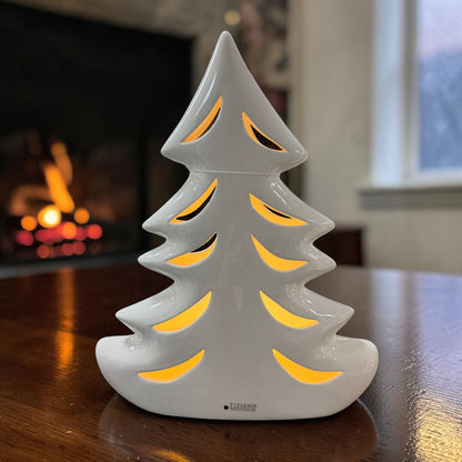 Cremeweißer Tiziano Keramik Tannenbaum mit warm beleuchteten Durchbrüchen auf einem Holztisch vor einem Kamin.