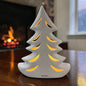 Cremeweißer Tiziano Keramik Tannenbaum mit warm beleuchteten Durchbrüchen auf einem Holztisch vor einem Kamin.