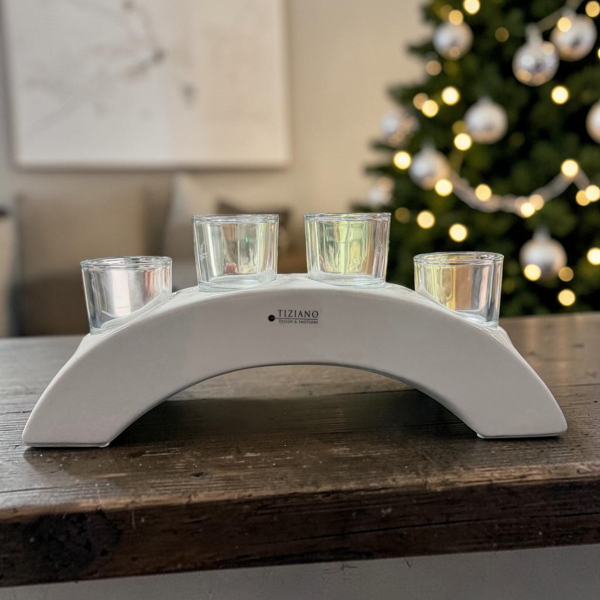 Cremeweißer Tiziano Keramik Teelichthalter in Bogenform mit vier Gläsern auf einem Holztisch vor beleuchtetem Weihnachtsbaum.