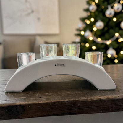 Cremeweißer Tiziano Keramik Teelichthalter in Bogenform mit vier Gläsern auf einem Holztisch vor beleuchtetem Weihnachtsbaum.