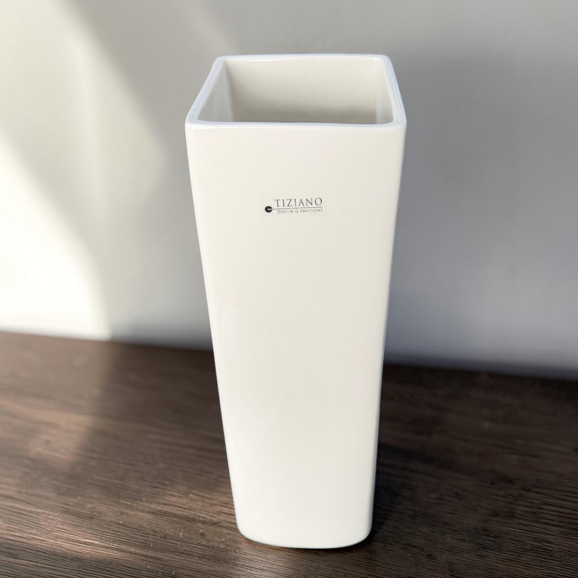 Hohe weiße Tiziano Keramik-Vase mit rechteckiger Öffnung auf einem dunklen Holztisch vor einer grauen Wand.