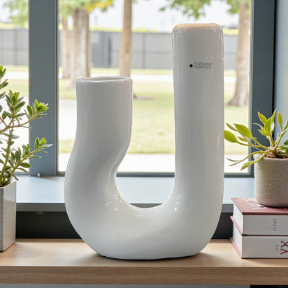 Keramik geschwungene U-Vase steht auf einer Fensterbank und ist von Pflanzen umgeben; ihre glänzende weiße Keramik wirkt modern und elegant im natürlichen Tageslicht.