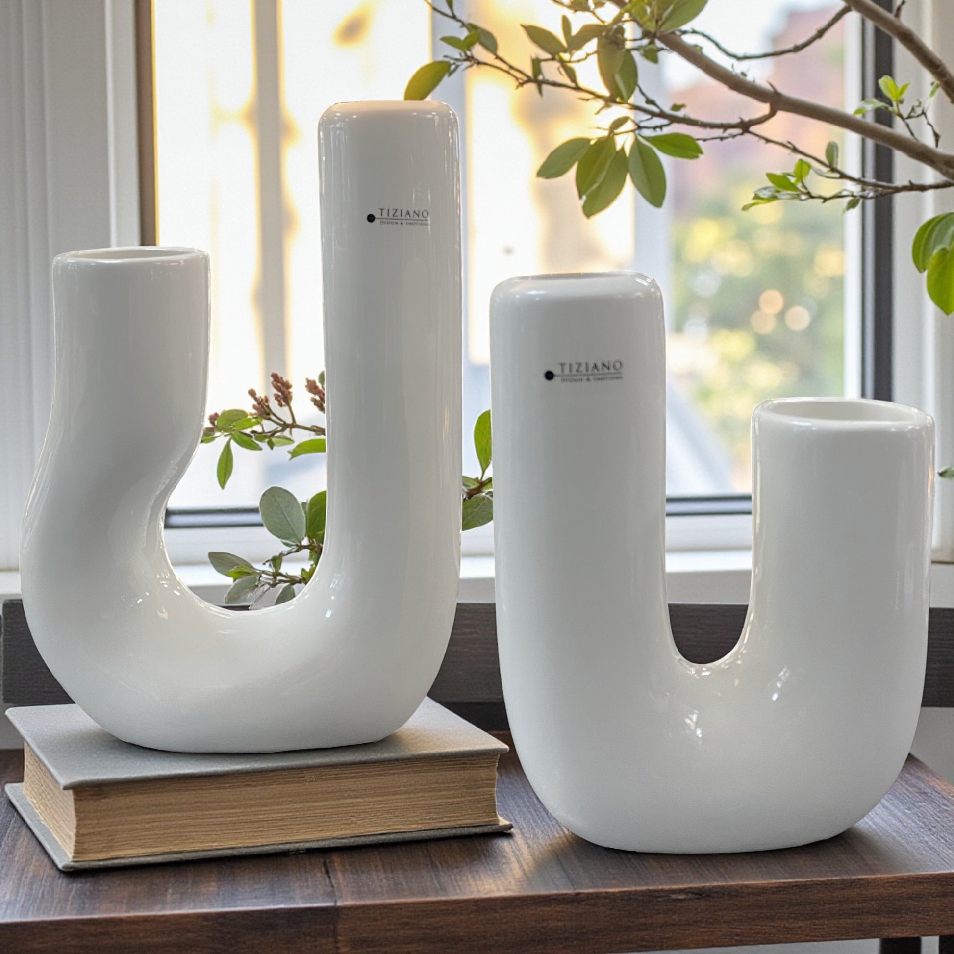 Zwei moderne Keramikvasen stehen als Dekoration auf einem Tisch vor einem Fenster, begleitet von einem Zweig; die matte weiße Oberfläche reflektiert das natürliche Licht.