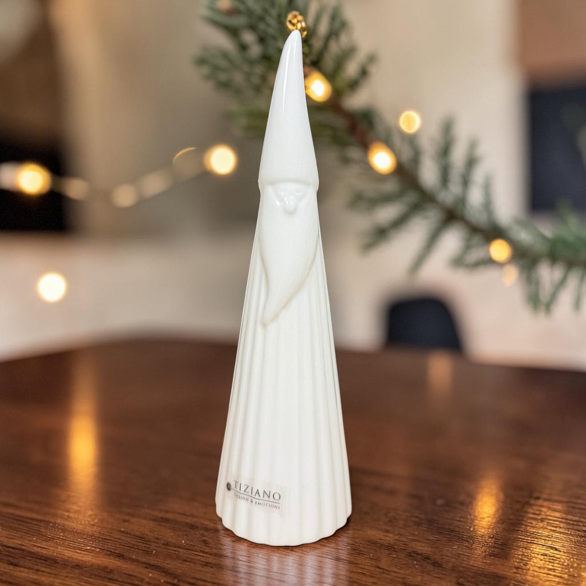 Hoher cremeweißer Tiziano Keramik-Weihnachtsmann mit langer Zipfelmütze auf einem Holztisch vor warm beleuchtetem weihnachtlichem Hintergrund.