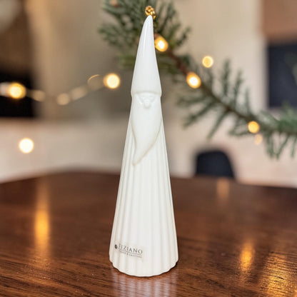 Hoher cremeweißer Tiziano Keramik-Weihnachtsmann mit langer Zipfelmütze auf einem Holztisch vor warm beleuchtetem weihnachtlichem Hintergrund.