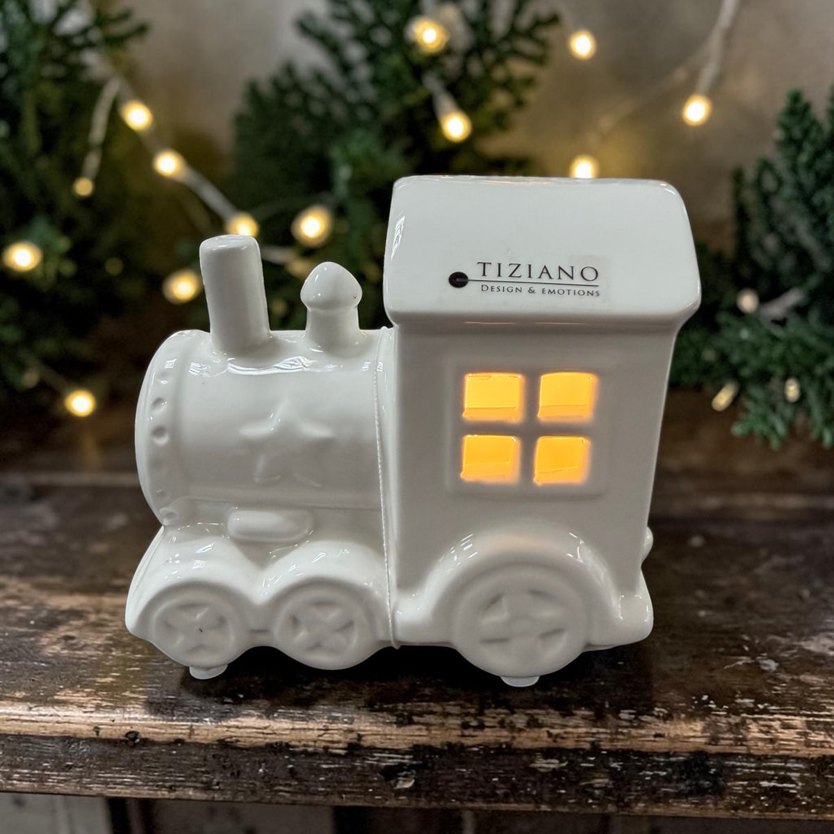 Cremeweiße Tiziano Keramik Lokomotive mit warm leuchtenden Fenstern auf einem dunklen Holztisch vor Tannenzweigen und Lichterkette.