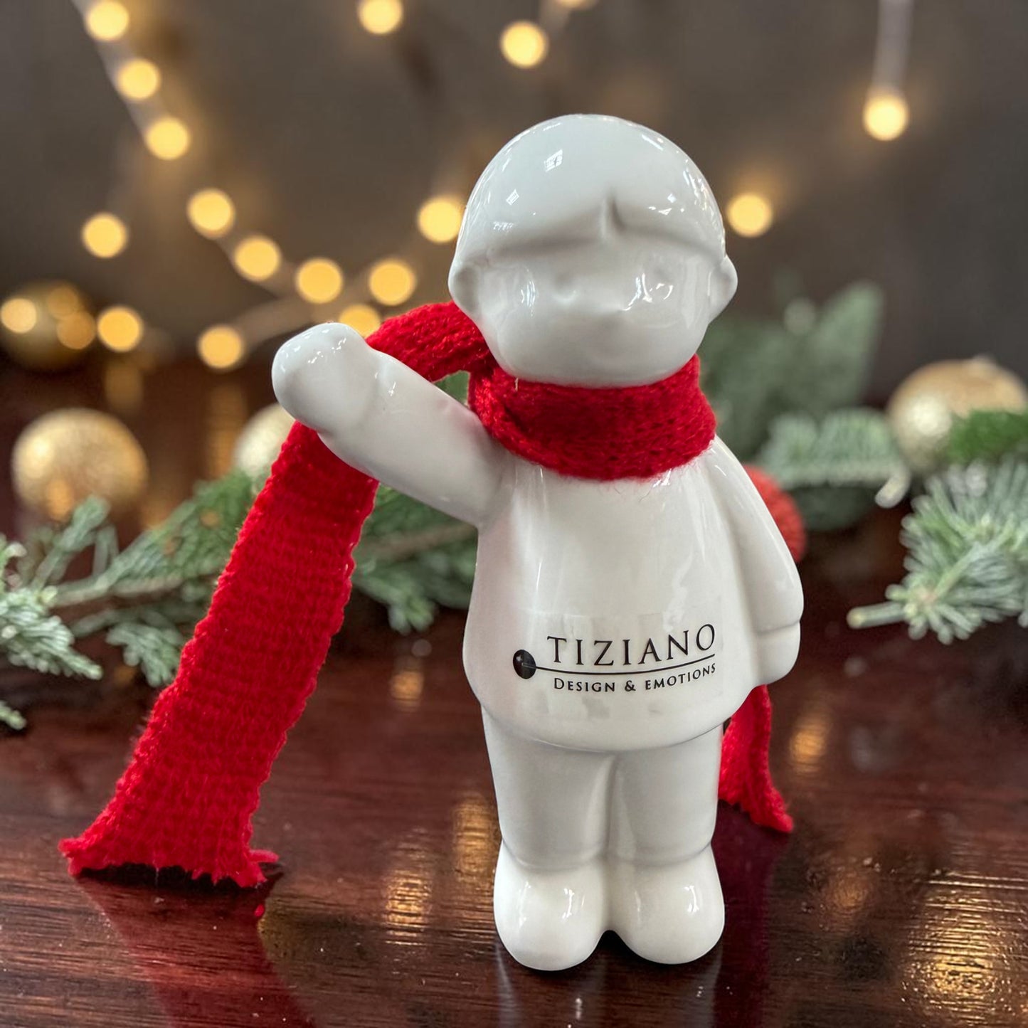 Cremeweißes Tiziano Keramik-Winterkind mit rotem Schal und erhobenem Arm auf einem dunklen Holztisch vor warmen Lichtern und Tannengrün.