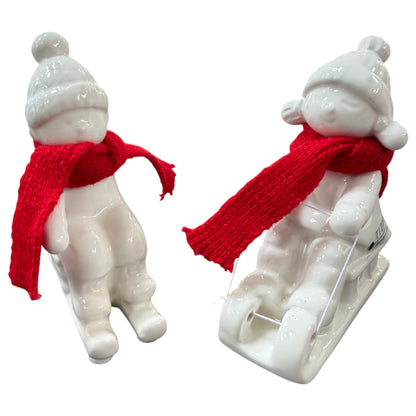 Zwei cremeweiße Tiziano Keramik-Winterkinder mit roten Schals, eines sitzend und eines auf einem Schlitten, vor weißem Hintergrund.