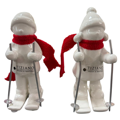 Zwei cremeweiße Tiziano Keramik-Winterkinder mit roten Schals und silberfarbenen Skistöcken vor weißem Hintergrund.