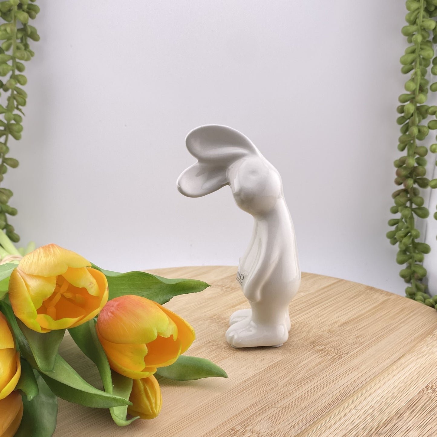 Ein kleiner creme-weißer Keramik Osterhase, der sein Gesicht zur Seite geneigt hat und mit seinen abstehenden Öhrchen lauschht. Er steht auf seinen Beinen und seine Arme hängen hinterunter. Der Deko Hase ist 13 cm hoch und im Ambiente abgebildet.