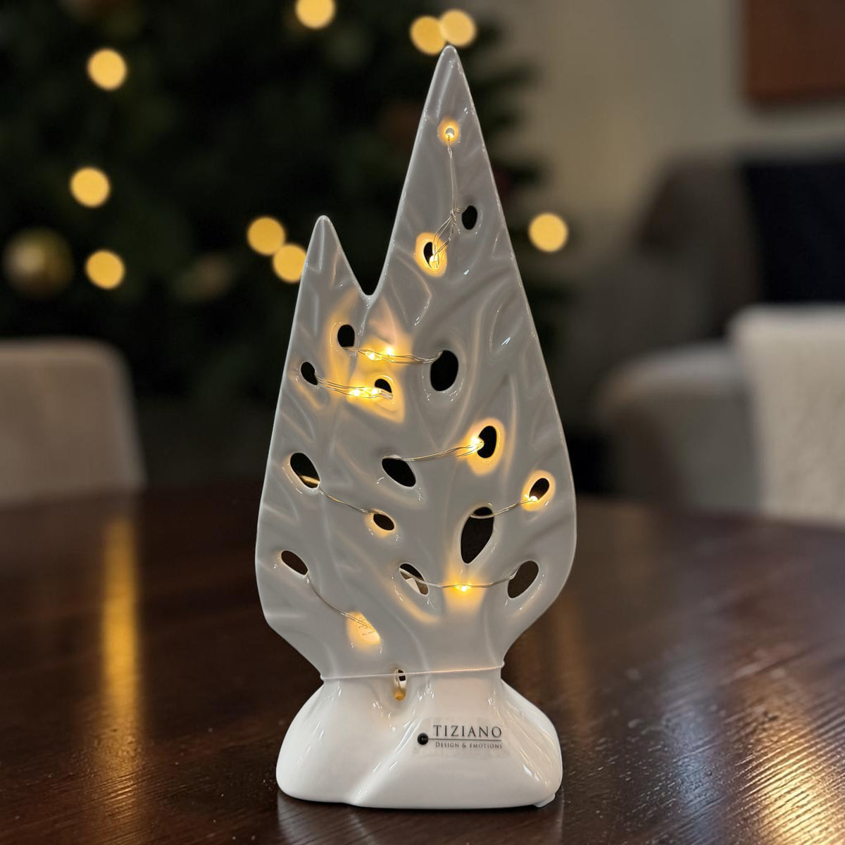 Cremeweißer Tiziano Keramik Deko Baum mit integrierter warmweißer Lichterkette auf einem Holztisch vor weihnachtlichem Hintergrund.