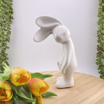 Ein creme-weißer Keramik Osterhase, der sein Gesicht zur Seite geneigt hat und mit seinen abstehenden Öhrchen lauschht. Er steht auf seinen Beinen und seine Arme hängen hinterunter. Der Deko Hase ist 18 cm hoch und im Ambiente abgebildet.