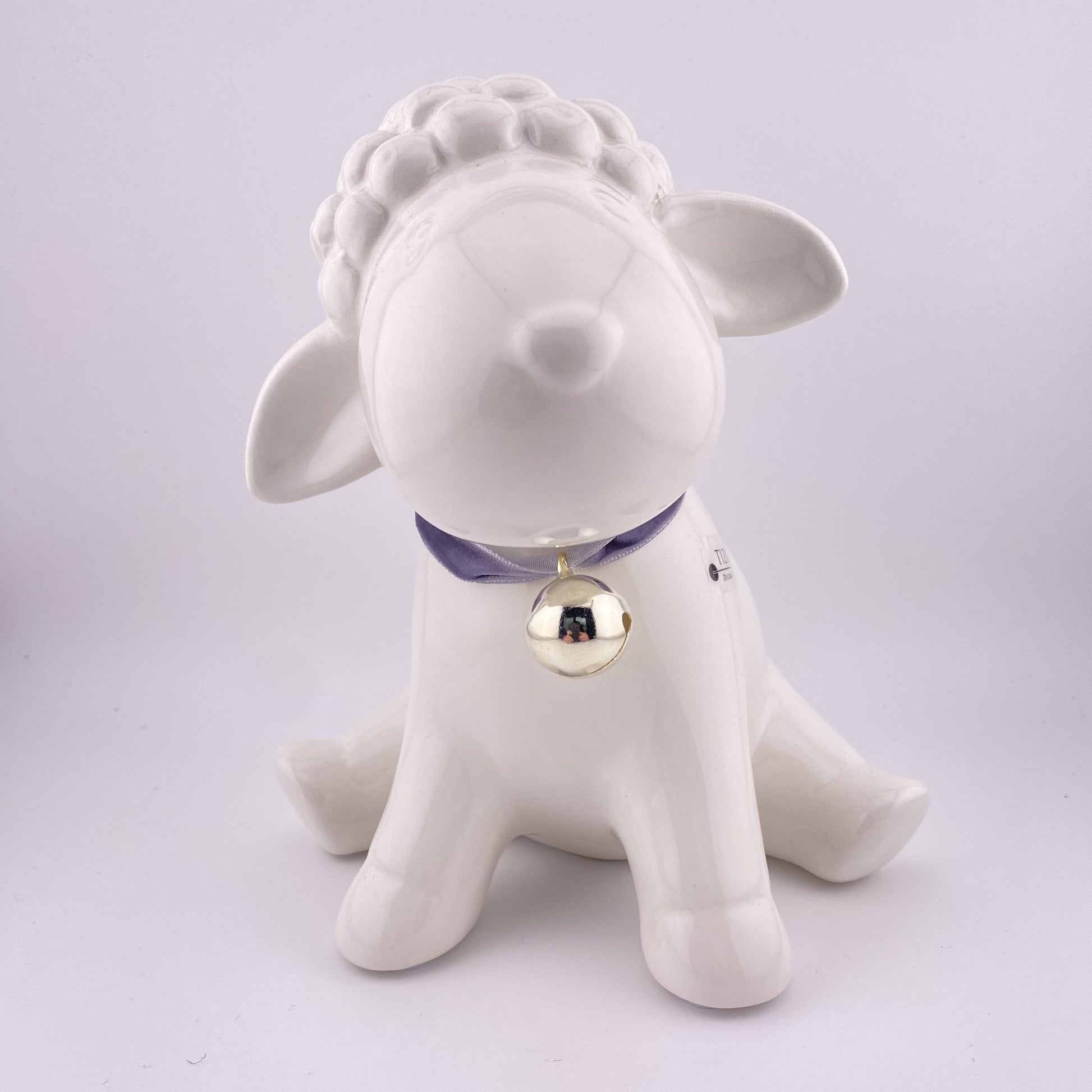 Ein großes Oster Deko Lamm aus creme-weißer Keramikmit gespitzten Ohren und Glocken-Halsband, das 17cm groß und vor weißem Hintergrund platziert ist. 