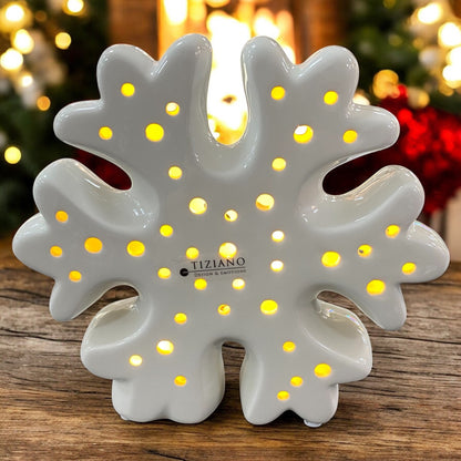 Eine weiße Schneeflocke aus Keramik mit kleinen runden Öffnungen und integrierter LED-Beleuchtung im Ambiente. Auf der Vorderseite ist das Logo „Tiziano Design & Emotions“ zu sehen.
