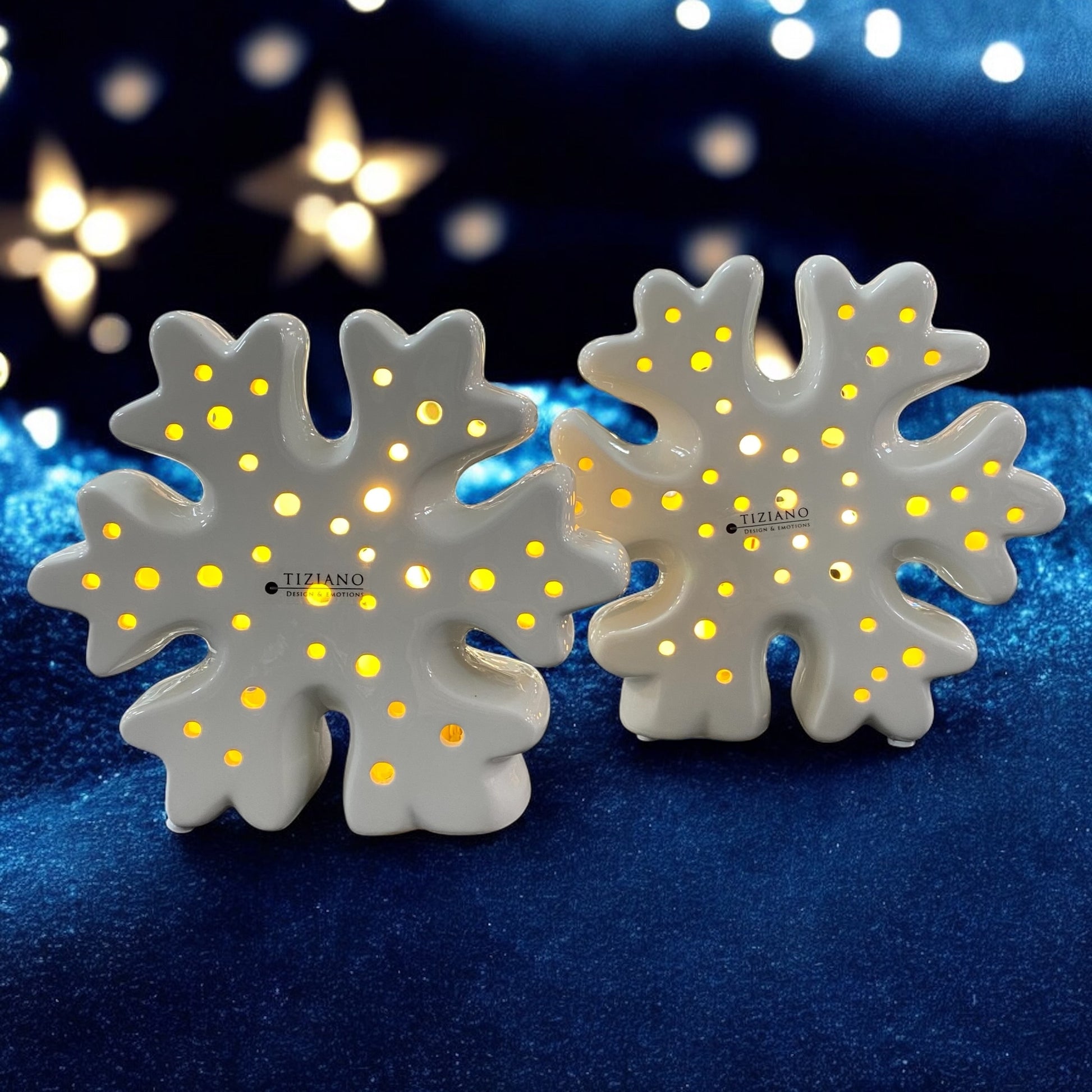 Zwei weiße Schneeflocken aus Keramik mit kleinen runden Öffnungen und integrierter LED-Beleuchtung im Ambiente. Die dekorativen Schneeflocken stehen nebeneinander und leuchten warmweiß. Auf der Vorderseite ist das Logo „Tiziano Design & Emotions“ zu sehen.