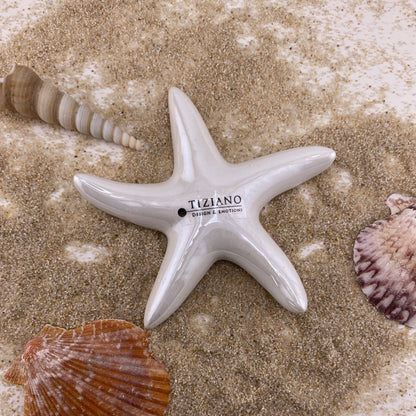 Ein Deko Objekt aus Keramik in Form eines Seesterns ist auf einem Teller mit Sand und Muscheln platziert. Der Seestern hat eine glatte Oberfläche in creme-weißer Farbe und ist 11cm groß.