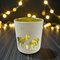 Cremefarbenes Teelichtglas mit Hirschmotiv und goldener Innenbeschichtung, beleuchtet vor unscharfem Lichterhintergrund.