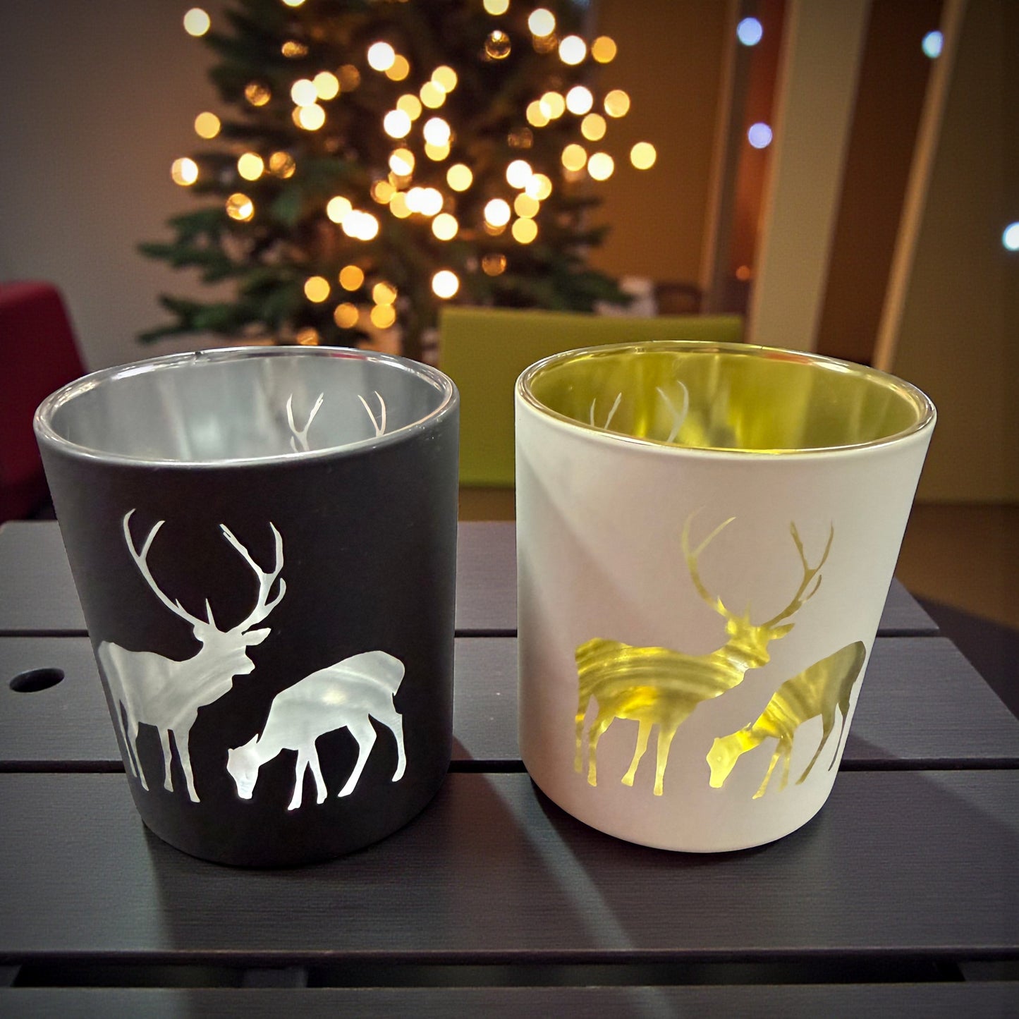 Teelichtgläser in Schwarz und Creme mit Hirschmotiv und goldener bzw. silberner Innenbeschichtung auf Tisch vor Weihnachtsbaum.