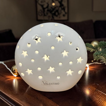 Beleuchtete Valentino Keramik Lichtkugel in Cremeweiß mit Sternen und runden Lichtöffnungen auf einem Holztisch vor weihnachtlicher Dekoration.