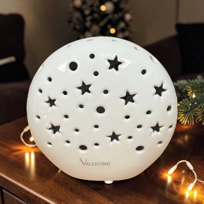 Valentino Keramik Lichtkugel in Cremeweiß mit sternförmigen und runden Aussparungen auf einem dunklen Holztisch mit weihnachtlicher Dekoration.