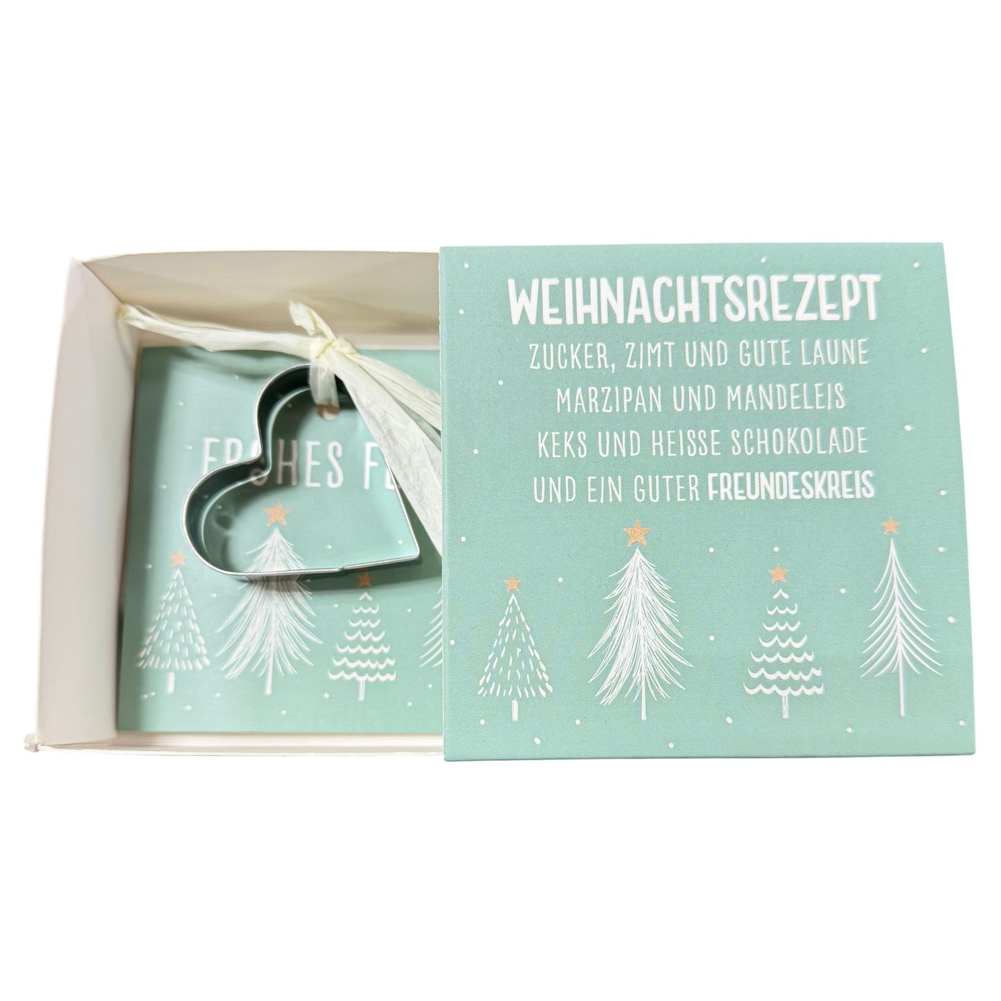 Offene Schachtel mit grüner Karte mit Aufschrift „Weihnachtsrezept“ und daran befestigter herzförmiger Ausstechform liegt vor weißem Hintergrund.