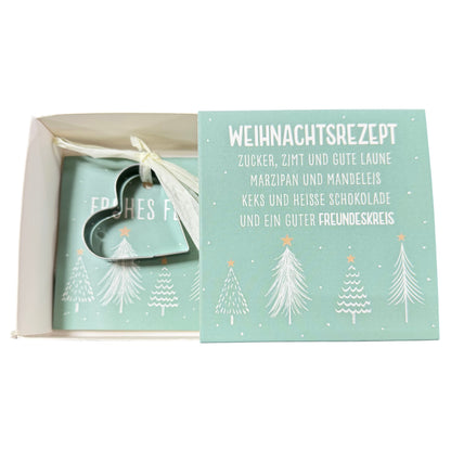 Offene Schachtel mit grüner Karte mit Aufschrift „Weihnachtsrezept“ und daran befestigter herzförmiger Ausstechform liegt vor weißem Hintergrund.