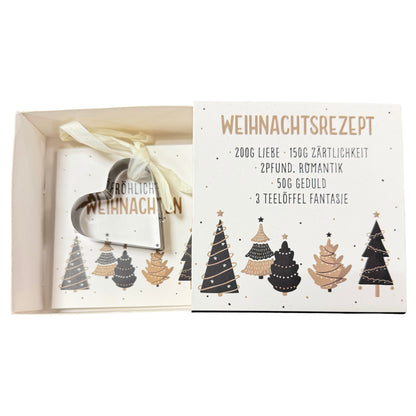 Offene Schachtel mit weißer Karte mit Aufschrift „Weihnachtsrezept“ und daran befestigter herzförmiger Ausstechform liegt vor weißem Hintergrund.